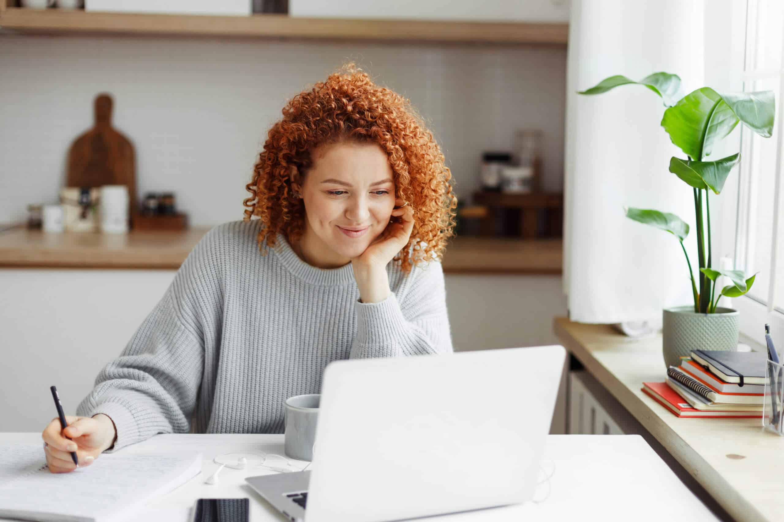 Cute girl with red curls watching online tutorial or lesson on laptop, smiling listening and looking attentively, making notes in copybook, sitting at kitchen table with smartphone and cup of coffee